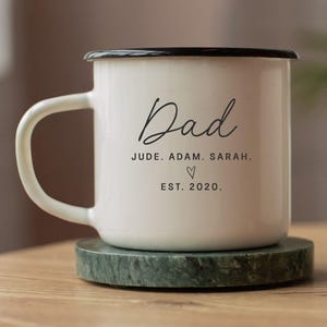May include: A white enamel camp mug with a black rim and handle. The mug features the word "Dad" in a script font, along with names and the year 2020. The mug is 10 oz and sits on a green marble coaster.
