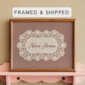 May include: A rectangular wooden frame containing a mauve fabric background with a white lace border surrounding the text Nora James in a cursive font. Above the frame, a white rectangular label displays the text FRAMED & SHIPPED.