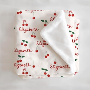 Puede incluir: Una manta para bebé blanca y crema con un patrón de cerezas rojas. La manta tiene el nombre "Elizabeth" impreso en ella varias veces. Un lado de la manta es suave y peludo.
