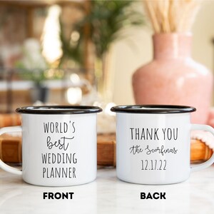 K&ouml;nnte beinhalten: Zwei wei&szlig;e Emaille-Tassen mit schwarzen R&auml;ndern und Henkeln. Auf einer Tasse steht in schwarzer Schrift "WORLD'S BEST WEDDING PLANNER". Auf der anderen Tasse steht "THANK YOU the Scorfinas 12.17.22".
