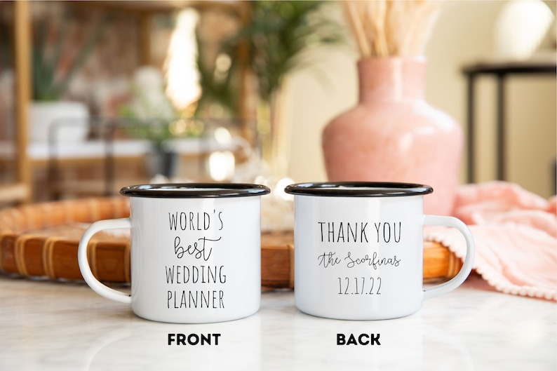 K&ouml;nnte beinhalten: Zwei wei&szlig;e Emaille-Tassen mit schwarzen R&auml;ndern und Henkeln. Auf einer Tasse steht in schwarzer Schrift "WORLD'S BEST WEDDING PLANNER". Auf der anderen Tasse steht "THANK YOU the Scorfinas 12.17.22".