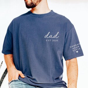 May include: A blue t-shirt with the word "dad" in cursive and "EST 2025" printed on the front. The sleeve features the names "HANNAH ADAM SOFIA" and a heart symbol. The shirt is a solid color and has a relaxed fit.