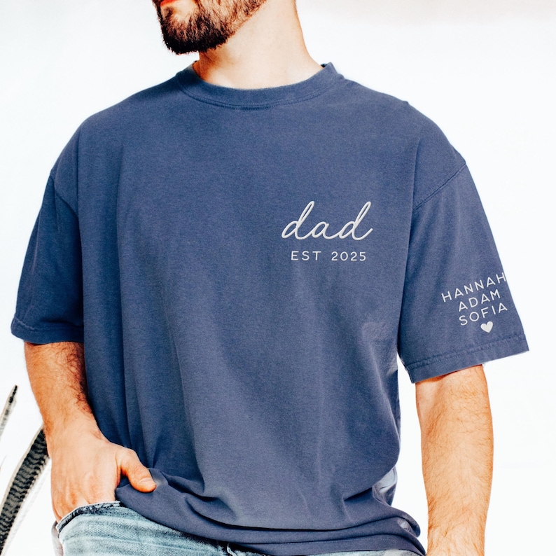 May include: A blue t-shirt with the word "dad" in cursive and "EST 2025" printed on the front. The sleeve features the names "HANNAH ADAM SOFIA" and a heart symbol. The shirt is a solid color and has a relaxed fit.