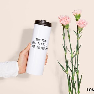 Peut inclure: Un long gobelet blanc avec un couvercle noir est tenu. Le gobelet porte le texte "CREATE YOUR MUG, PICK TEXT, FONT, AND DESIGN". Des &oelig;illets roses avec des tiges vertes sont &agrave; droite. Le mot "LONG TUMBLER" est en bas.