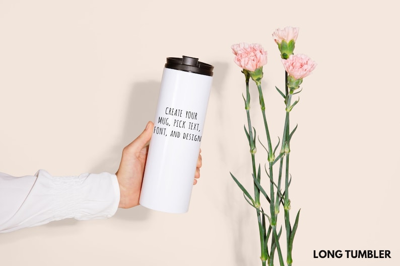 Peut inclure: Un long gobelet blanc avec un couvercle noir est tenu. Le gobelet porte le texte "CREATE YOUR MUG, PICK TEXT, FONT, AND DESIGN". Des &oelig;illets roses avec des tiges vertes sont &agrave; droite. Le mot "LONG TUMBLER" est en bas.