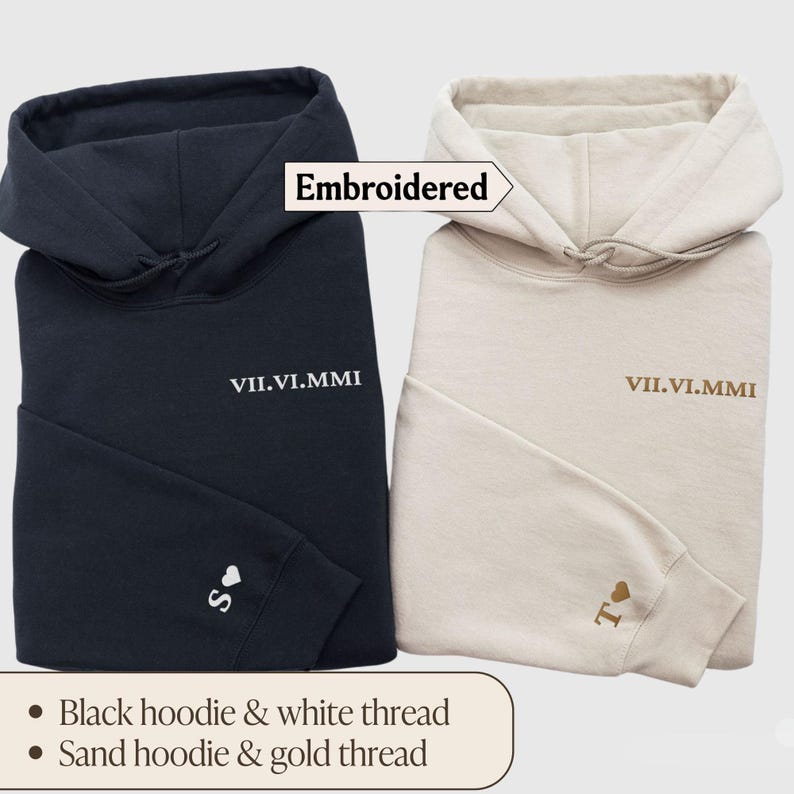 May include: Two hooded sweatshirts, one black and one sand colored, with embroidered text "VII.VI.MMI" and a small heart with a letter on each. The black hoodie has white thread embroidery and the sand hoodie has gold thread embroidery.