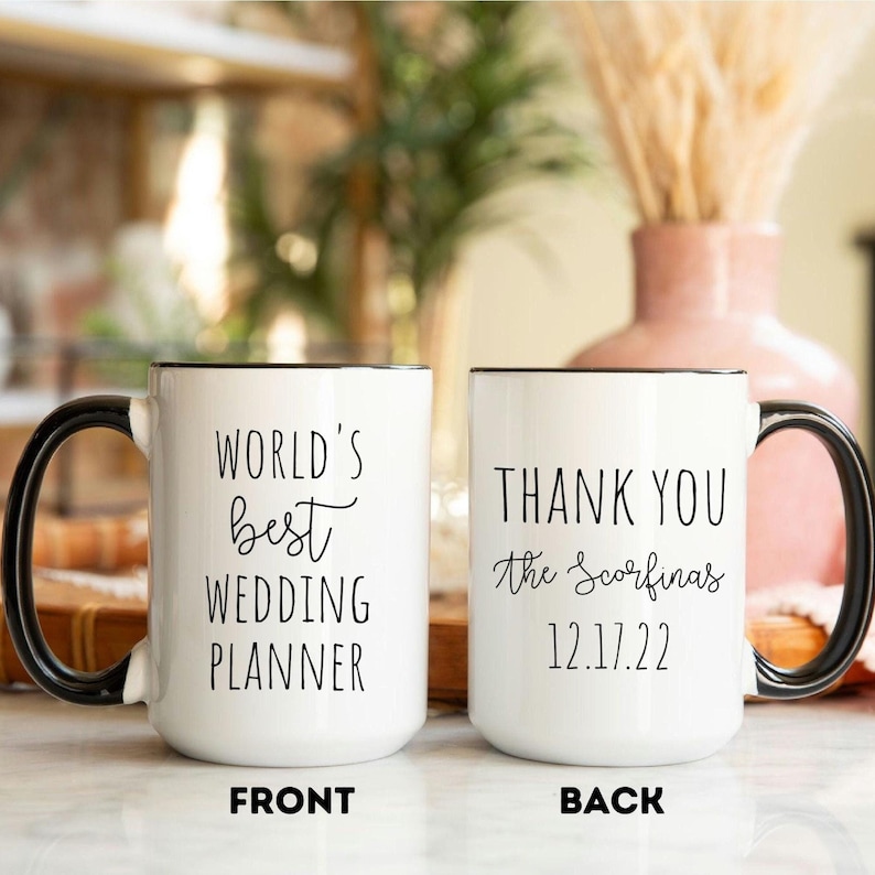K&ouml;nnte beinhalten: Wei&szlig;e Keramiktasse mit schwarzem Griff und Rand. Auf der Vorderseite der Tasse steht "WORLD'S best WEDDING PLANNER". Auf der R&uuml;ckseite der Tasse steht "THANK YOU the Scorfina's 12.17.22".