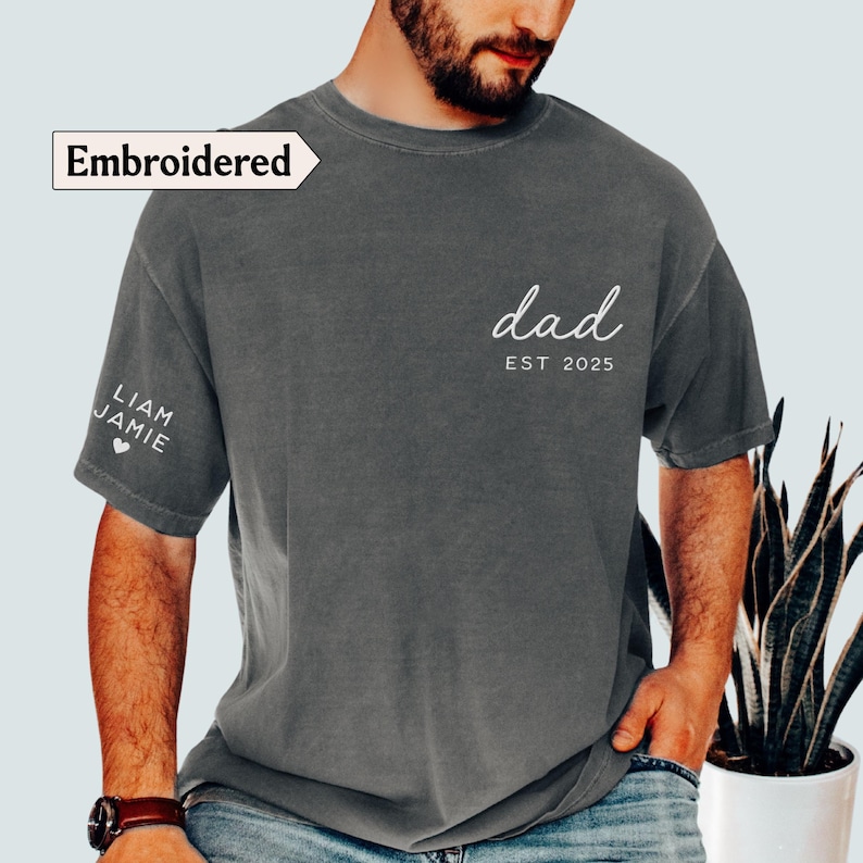 May include: A dark gray t-shirt with the word "dad" and "EST 2025" printed in white script on the front. The left sleeve has the names "LIAM JAMIE" and a heart symbol. The word "Embroidered" is in a white box.