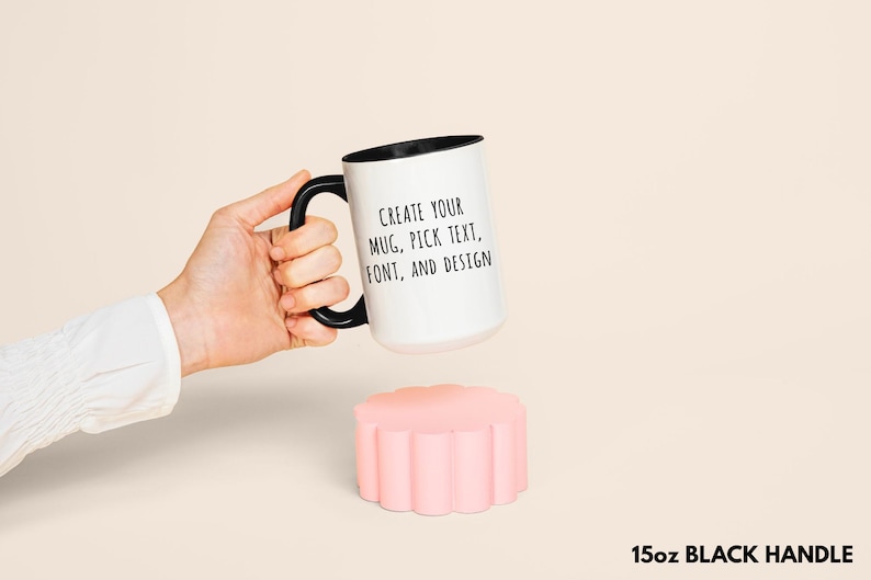 Peut inclure: Mug blanc avec anse et bord noir, avec le texte "CREATE YOUR MUG, PICK TEXT, FONT, AND DESIGN." Le mug est tenu au-dessus d'un dessous de verre rose &agrave; bords festonn&eacute;s. Le mug est de 15 oz.