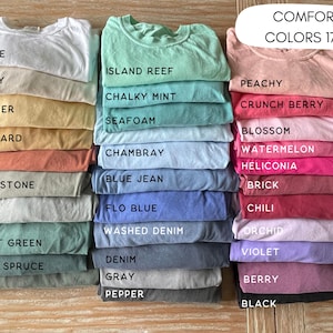 May include: Stacks of folded t-shirts in various colors, including white, ivory, butter, and mustard. The image also displays shirts in shades of blue, green, pink, and gray. The text "COMFORT COLORS 1717" is visible.