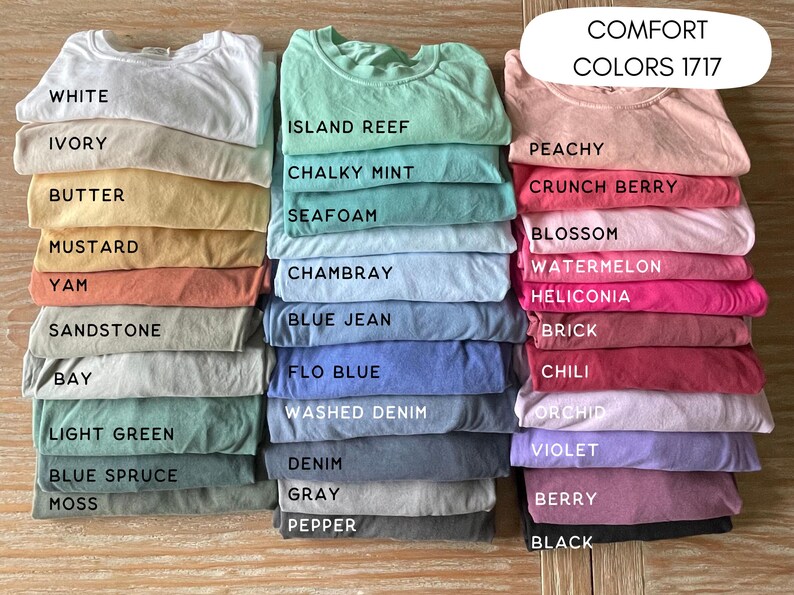 May include: Stacks of folded t-shirts in various colors, including white, ivory, butter, and mustard. The image also displays shirts in shades of blue, green, pink, and gray. The text "COMFORT COLORS 1717" is visible.