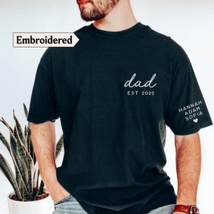 May include: Black t-shirt with the word "dad" in white script, the year "EST 2025", and names on the sleeve. The shirt has the word "Embroidered" in a white box. The shirt is a dark color.