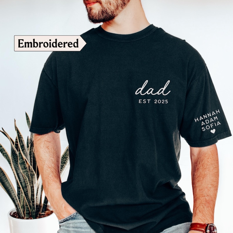 May include: Black t-shirt with the word "dad" in white script, the year "EST 2025", and names on the sleeve. The shirt has the word "Embroidered" in a white box. The shirt is a dark color.