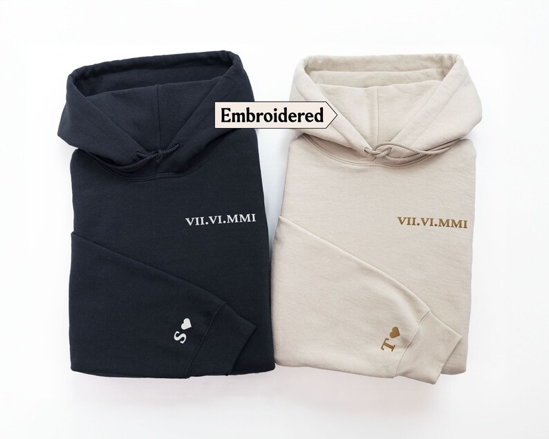 May include: Two hooded sweatshirts, one black and one beige, with embroidered text and a heart on the sleeve. The text on both sweatshirts reads "VII.VI.MMI". The heart on the black sweatshirt is white and the heart on the beige sweatshirt is gold.