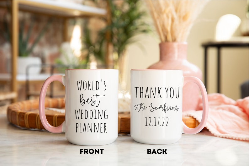 K&ouml;nnte beinhalten: Wei&szlig;er Keramikbecher mit rosa Griff und Rand. Auf der Vorderseite des Bechers steht "WORLD'S best WEDDING PLANNER" in schwarzer Schrift. Auf der R&uuml;ckseite des Bechers steht "THANK YOU the Scorfina's 12.17.22".