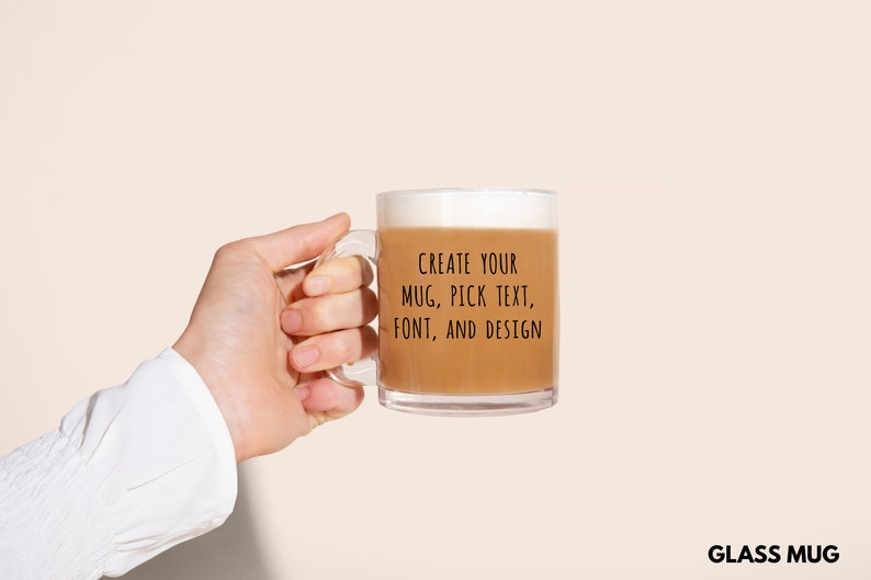 Peut inclure: Un mug en verre transparent rempli d'une boisson brun clair, tenu par une personne. Le mug a une anse et le texte "CREATE YOUR MUG, PICK TEXT, FONT, AND DESIGN." Les mots "GLASS MUG" sont en bas.
