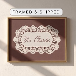 May include: A rectangular wall art piece in a gold frame featuring a dark maroon background. Centered is an off-white lace doily graphic with a scalloped edge. The text "The Clarks" is printed in a dark maroon cursive font inside the lace border.