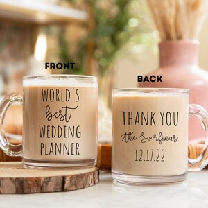 K&ouml;nnte beinhalten: Klarer Glasbecher gef&uuml;llt mit einem cremigen Getr&auml;nk. Auf der Vorderseite des Bechers steht "WORLD'S best WEDDING PLANNER" in schwarzer Schrift. Auf der R&uuml;ckseite steht "THANK YOU the Scorfina's 12.17.22". Der Becher steht auf einer Holzscheibe.