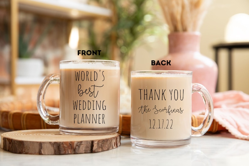 K&ouml;nnte beinhalten: Klarer Glasbecher gef&uuml;llt mit einem cremigen Getr&auml;nk. Auf der Vorderseite des Bechers steht "WORLD'S best WEDDING PLANNER" in schwarzer Schrift. Auf der R&uuml;ckseite steht "THANK YOU the Scorfina's 12.17.22". Der Becher steht auf einer Holzscheibe.