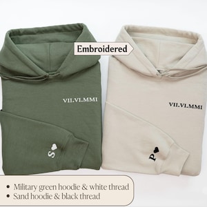 May include: Two embroidered hoodies, one in military green with white thread and the other in sand with black thread. Both hoodies feature the text "VII.VI.MMI" and a small heart on the sleeve.