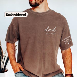 May include: Brown t-shirt with the word "dad" and "EST 2025" printed in white. The sleeve features the names "HANNAH ADAM SOFIA" and a heart symbol. The shirt has the word "Embroidered" on the top left.