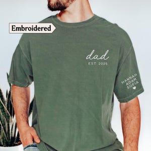 May include: Olive green t-shirt with the word "dad" and "EST 2025" in white script. The sleeve features the names "Hannah, Adam, Sofia" and a heart. The shirt has the word "Embroidered" in a white box.