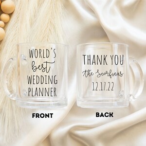 K&ouml;nnte beinhalten: Zwei transparente Glasbecher mit Henkeln. Auf einem Becher steht "WORLD'S best WEDDING PLANNER" und auf dem anderen "THANK YOU the Scorfina's 12.17.22". Die Becher stehen auf einer cremefarbenen Satin-Oberfl&auml;che.