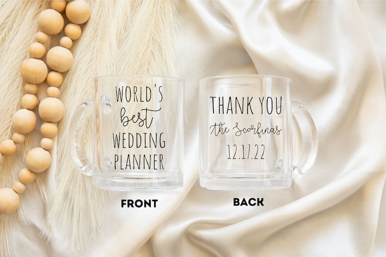 K&ouml;nnte beinhalten: Zwei transparente Glasbecher mit Henkeln. Auf einem Becher steht "WORLD'S best WEDDING PLANNER" und auf dem anderen "THANK YOU the Scorfina's 12.17.22". Die Becher stehen auf einer cremefarbenen Satin-Oberfl&auml;che.