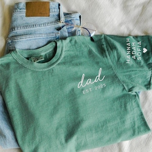 May include: A sage green t-shirt with the word "dad" in white script and "EST 2025" printed below. The sleeve features names and a heart symbol. Light blue jeans and eyeglasses are also in the image.