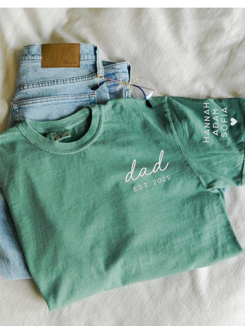 May include: A sage green t-shirt with the word "dad" in white script and "EST 2025" printed below. The sleeve features names and a heart symbol. Light blue jeans and eyeglasses are also in the image.