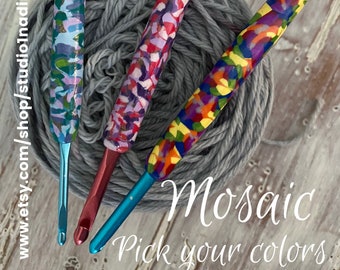 May include: Three mosaic-patterned crochet hooks in shades of blue, purple, and rainbow colors, resting on a gray ball of yarn. The hooks have metallic shafts and are labeled "Mosaic Pick your colors."