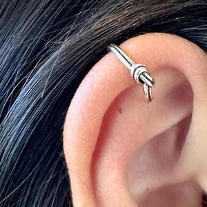 May include: A silver knot ear cuff. The cuff is a simple, thin band of metal with a knot in the center. The cuff is worn in the upper cartilage of the ear.