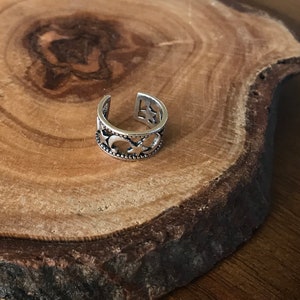 May include: Silver ear cuff with a delicate, intricate design featuring a repeating pattern of circles and stars. The cuff is displayed on a wooden surface.