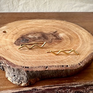 May include: Two gold-colored wavy line earrings on a wooden surface.