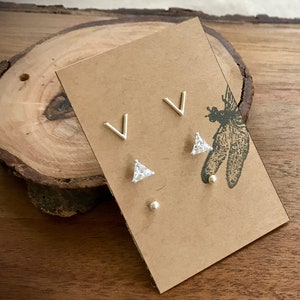 May include: A set of three silver earrings on a brown card. The earrings are a pair of small silver triangles with a clear stone in the center, and a pair of small silver V shapes.