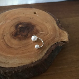May include: A silver earring with two white pearl accents. The earring is on a wooden surface.