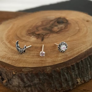 May include: Three silver nose rings on a wooden surface. The rings are a crescent moon, a small round gem, and a round ring with a white stone.