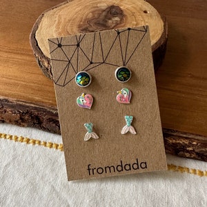 May include: A set of three pairs of stud earrings on a brown card with the text "fromdada". The earrings feature a mermaid tail, a heart with a unicorn, and a mermaid scale design.