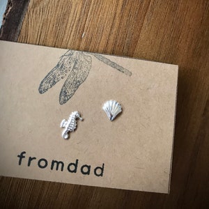May include: A pair of silver earrings featuring a seashell and a seahorse design. The earrings are displayed on a brown card with the text "fromdad" printed on it.