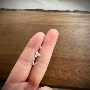 May include: A silver body piercing with a small, clear triangle-shaped gem and a small silver ball. The piercing is held in a hand.