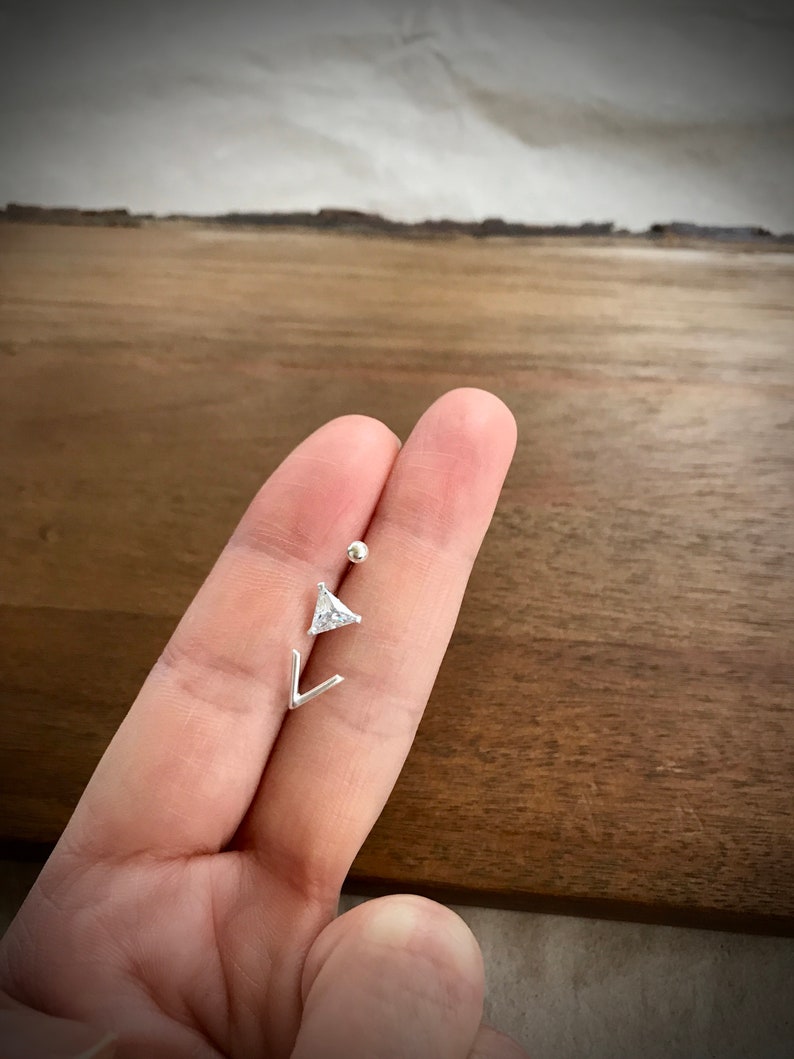 May include: A silver body piercing with a small, clear triangle-shaped gem and a small silver ball. The piercing is held in a hand.