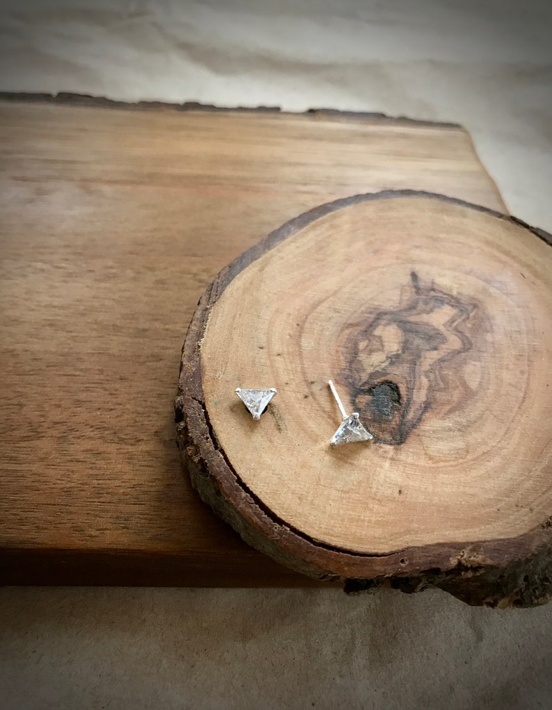 May include: A pair of silver triangle stud earrings with clear cubic zirconia stones on a wooden background.