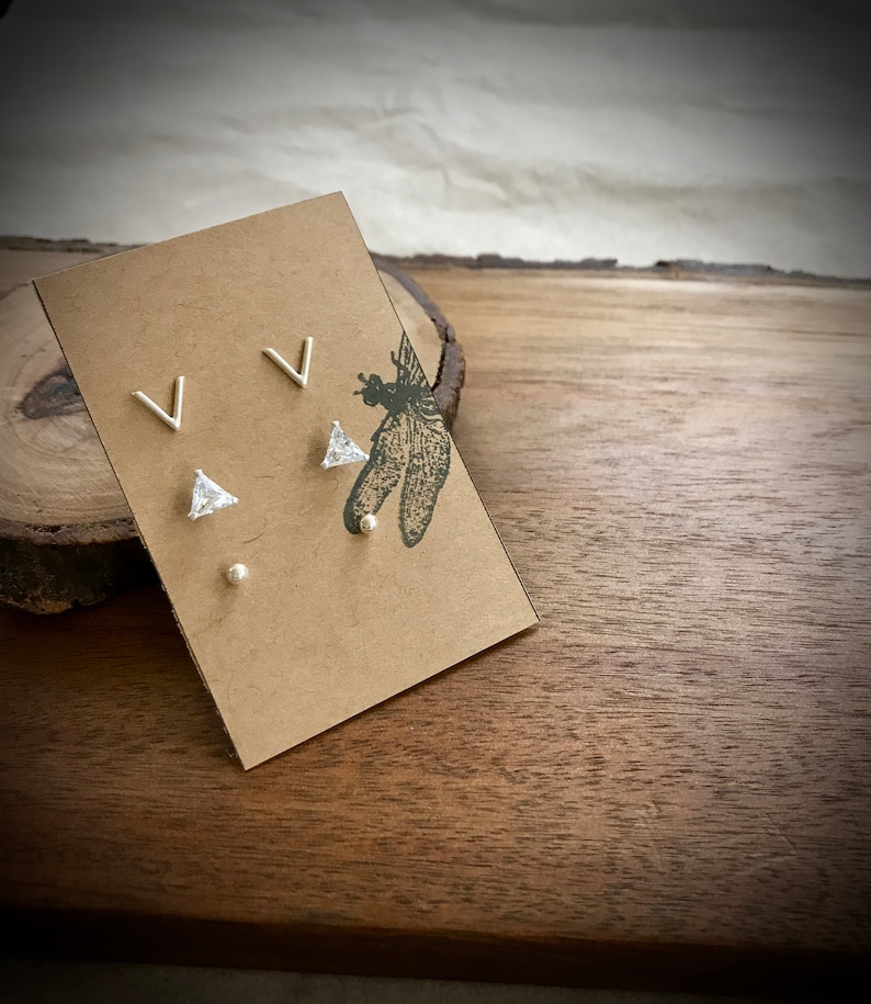May include: A set of three silver stud earrings on a brown card. The earrings are a small triangle with a clear stone, a small silver ball, and a silver V shape.