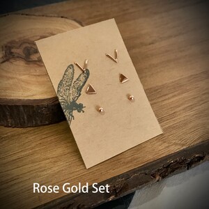 May include: A set of rose gold earrings featuring a triangle, a V shape, and a small round stud. The earrings are displayed on a brown card with the text "Rose Gold Set".