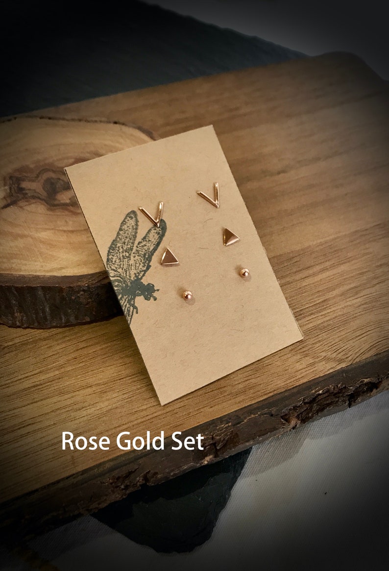 May include: A set of rose gold earrings featuring a triangle, a V shape, and a small round stud. The earrings are displayed on a brown card with the text "Rose Gold Set".
