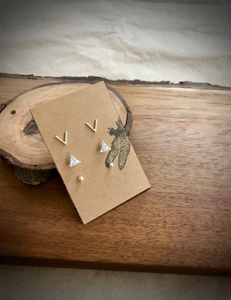 May include: A set of three silver earrings on a brown card. The earrings are a pair of small silver triangles with a clear stone in the center, and a pair of small silver V shapes.