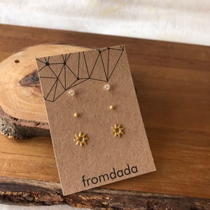 May include: A set of gold earrings featuring a small sun design and two small round studs. The earrings are displayed on a brown card with the text "fromdada" printed on it.
