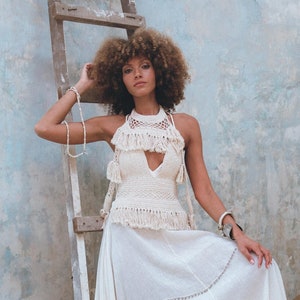 May include: A woman wearing a white crochet halter top with fringe and a white maxi skirt with fringe. She is standing in front of a blue wall with a wooden ladder behind her.