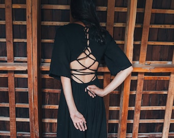 May include: A woman wearing a black dress with a lace-up back stands in front of a wooden wall.