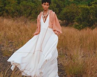 May include: A woman wearing a white maxi dress with a peach-colored cropped jacket and a shell necklace stands in a field of tall grass with a mountain in the background.